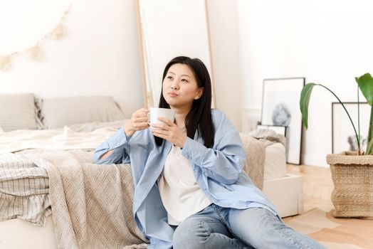 Photos of a charming young Asian woman sitting on the floor by the bed, drinking coffee and enjoying the weekend in the morning