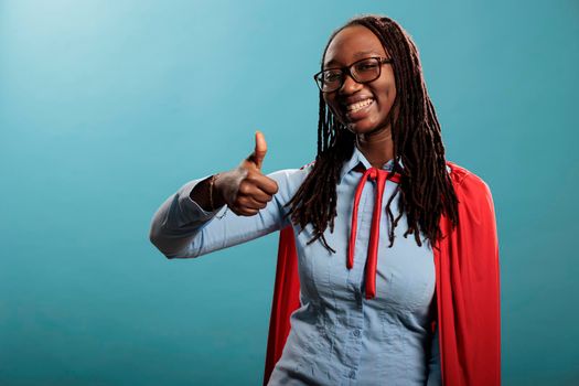 Young justice defender wearing red hero mighty cape showing thumbs up gesture sign at camera on blue background. Brave and proud superhero woman wearing cloak while smiling heartily. Studio shot.