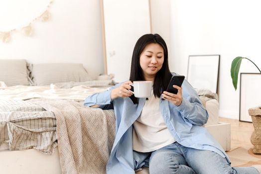 Photos of a charming young Asian woman sitting on the floor by the bed, drinking coffee and enjoying the weekend in the morning. A young Korean woman drinks warm tea, watching news on the phone