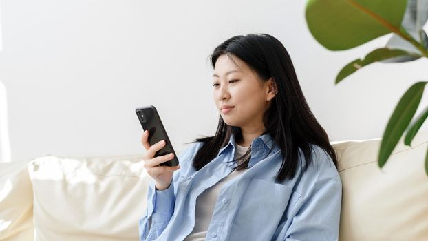 Young Asian woman relaxing on the sofa in the living room, browsing the news on her smartphone. Korean lady writes messages on modern mobile phone, shopping online through website. Web banner.