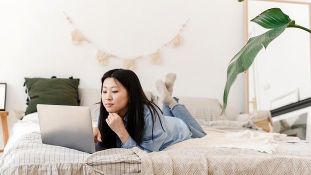 Beautiful calm asian female freelancer is working on a laptop while lying on the bed. Korean young woman doing online shopping, online training. Web banner.