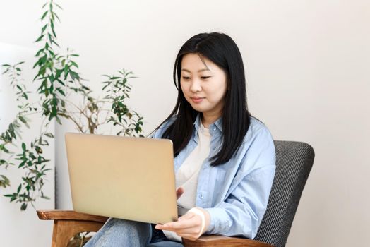 Freelance concept. Asian young woman sits in an armchair with a laptop working at home