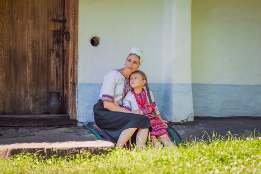 mother and daughter in Ukrainian national costumes are sitting near an old house