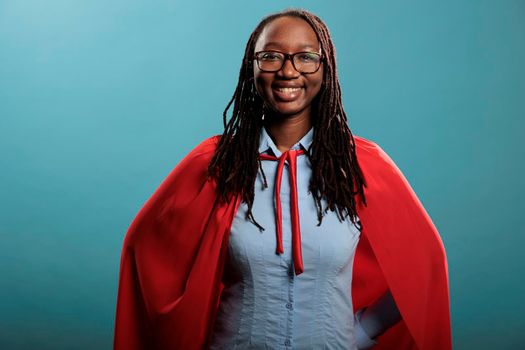 Smiling heartily young adult superhero woman wearing red hero cape while posing at camera. Portrait of brave and proud african american woman being happy and joyful on blue background. Studio shot.