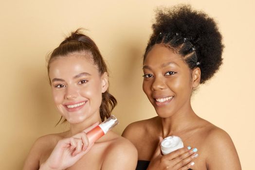 Smiling women hold jat of cosmetic moisturizer or face mask. Photo of attractive multiethnic women with perfect make-up on a beige background. Beauty concept.