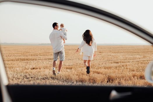 Young Family Walking in Field at Sunset, Back View Through Car Window Dad Carry His Daughter Having Weekend Time with Mom