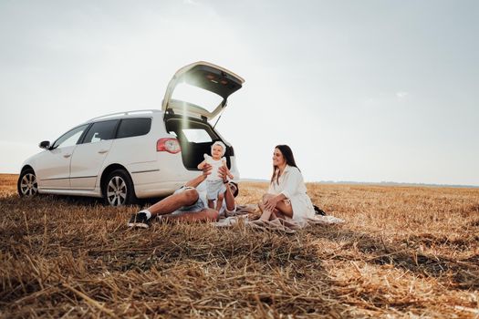 Conceptual Photo of Young Family Enjoying Their Road Trip with Car, Mom and Dad with Their Toddler Daughter Having Picnic Outdoors in Field at Sunset
