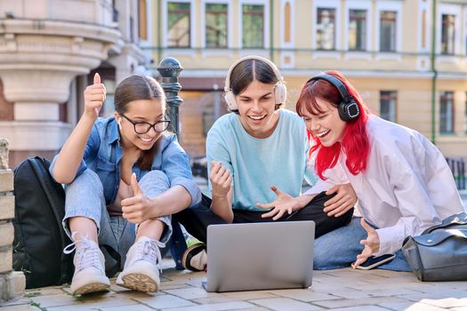 Teenage friends in headphones looking at laptop. Group of young people having fun laughing looking at laptop screen, sitting on sidewalk, urban style background. Youth, fun, lifestyle, leisure concept