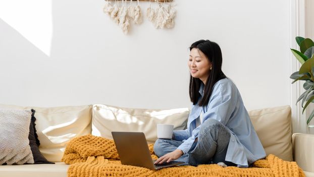 Portrait of smiling asian woman sitting on sofa using laptop in cozy living room at home. Happy female student using computer for leisure, study, education or work. Web banner.