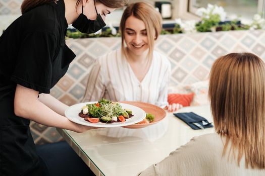 Female Waiter Bringing Order for Two Young Women in Restaurant