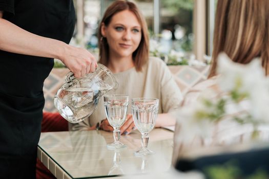 Close Up of Female Waiter Pours Water to Glass for Two Women in Restaurant