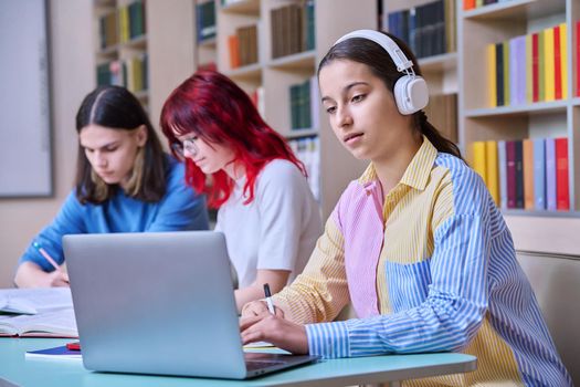 Group of teenage students study in school library classroom. In focus is teenager girl in headphones using laptop. High school, learning, education, knowledge, adolescence, people concept