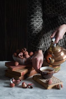 A woman pours tea from a copper teapot into a cup against the background of a stack of books and a cup with lychee, homely cozy tea drinking and reading,. High quality photo