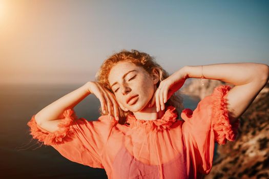 Side view a Young beautiful sensual woman in a red long dress posing on a volcanic rock high above the sea during sunset. Girl on the nature on overcast sky background. Fashion photo