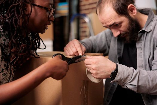 Diverse life partners using sticky tape and scissors to pack furniture in boxes, moving in new household property together. Packing things with adhesive roller to move in rented flat. Close up.