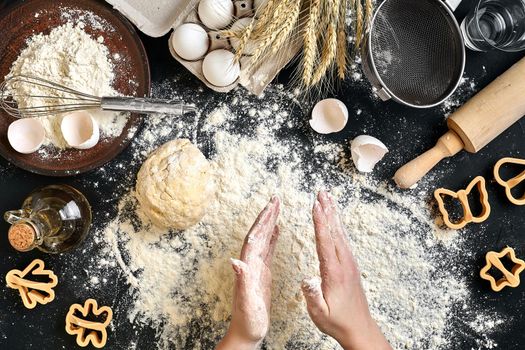 Woman's hands knead dough on table with flour, eggs and ingredients on black table. Top view. Still life. Flat lay