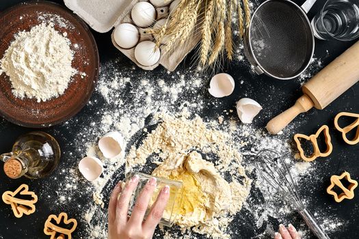 Woman's hands knead dough on table with flour, eggs and ingredients on black table. Top view. Still life. Flat lay