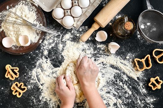 Woman's hands knead dough on table with flour, eggs and ingredients on black table. Top view. Still life. Flat lay
