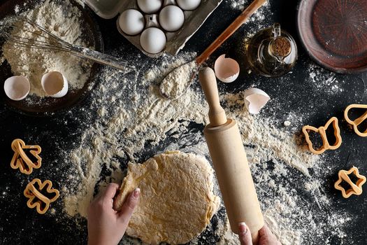 Female hands rolling dough with a rolling pin on a black table. Ingredients for the dough: flour, oil and eggs. Top view. Still life. Flat lay