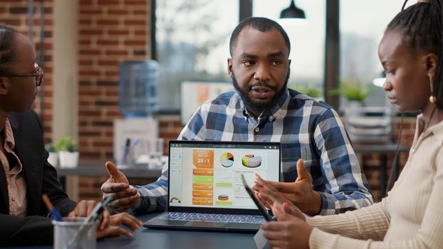 Team of african american people meeting to analyze sales charts and financial statistics on laptop display. Office workers collaborating on marketing presentation, brainstorming ideas.