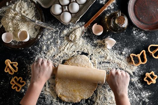 Female hands rolling dough with a rolling pin on a black table. Ingredients for the dough: flour, oil and eggs. Top view. Still life. Flat lay