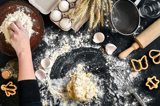 Woman's hands knead dough on table with flour, eggs and ingredients on black table. Top view. Still life. Flat lay