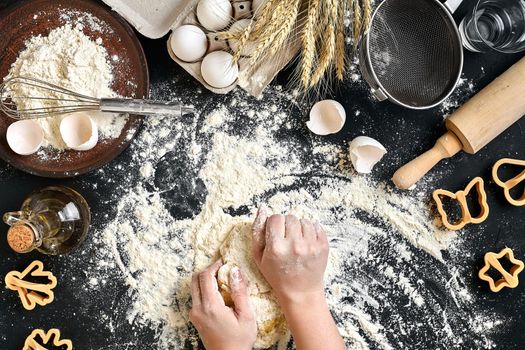 Woman's hands knead dough on table with flour, eggs and ingredients on black table. Top view. Still life. Flat lay