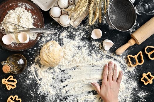 Woman's hands knead dough on table with flour, eggs and ingredients on black table. Top view. Still life. Flat lay