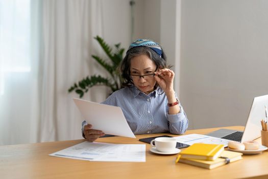 Seriously senior woman tworking at home, looking at laptop and waving paper correspondence, sitting at home, free space