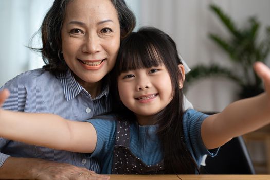 Close-up portrait of joyful happy elderly grandmother is taking selfie with her adorable granddaughter. They are looking at camera with wide smile at home