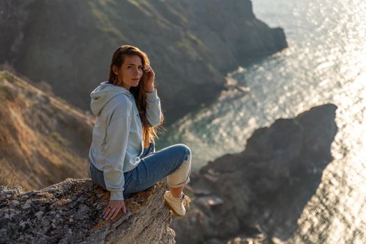 Woman tourist enjoying the sunset over the sea mountain landscape. Sits outdoors on a rock above the sea. She is wearing jeans and a blue hoodie. Healthy lifestyle, harmony and meditation.