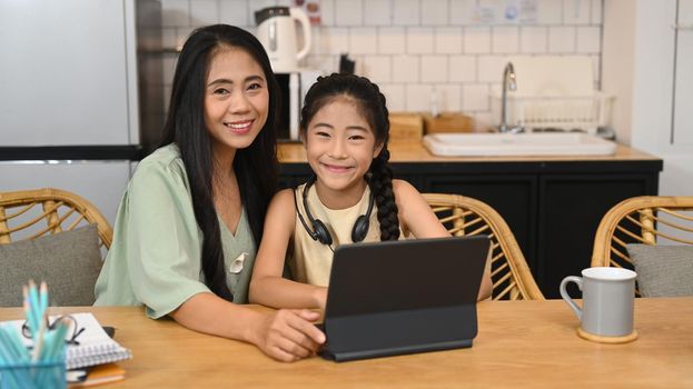Asian mother and little daughter sitting in kitchen, studying online, doing homework, learning at home, homeschooling concept.