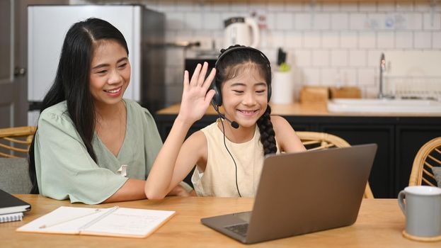 Cheerful asian girl in headphones talking to teacher while studying online via laptop at home with happy mom sitting nearby and giving support.
