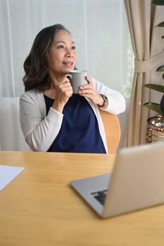 Relaxed middle aged woman holding cup of coffee, daydreaming while looking away. Retirement, technology concept.