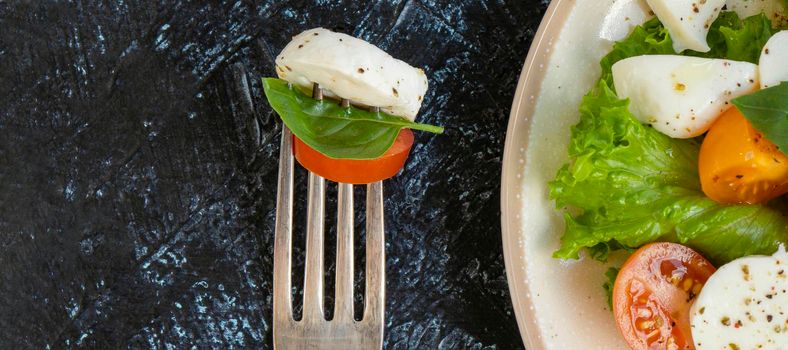 banner with Caprese salad with tomatoes, mozzarella, basil and olive oil on a black texture background. fork and plate with vegetable salad with cheese. Top view. Soft focus