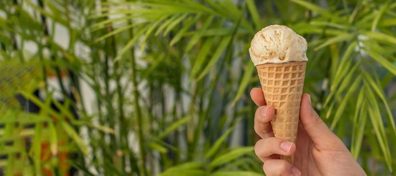 banner with young hand holds an ice cream cone on a background of green palm trees. the concept of relaxation by the sea on a bright, hot and sunny day on the beach. soft focus