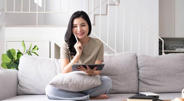 Beautiful smiling Asian woman looking at camera relaxing on leather sofa at home. work on a laptop computer.