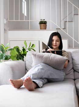 Attractive smiling young asian woman relaxing on a leather couch at home, working on laptop computer..