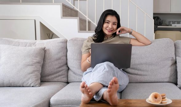 Beautiful smiling Asian woman looking at camera relaxing on leather sofa at home. work on a laptop computer.