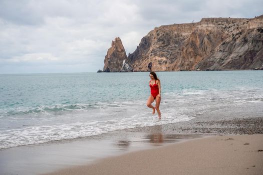 A beautiful and sexy brunette in a red swimsuit on a pebble beach, Running along the shore in the foam of the waves.