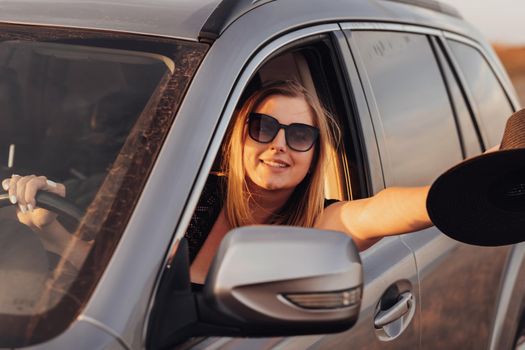 Cheerful Young Woman Driving Car and Showing Hat in Hand Through Window at Sunset