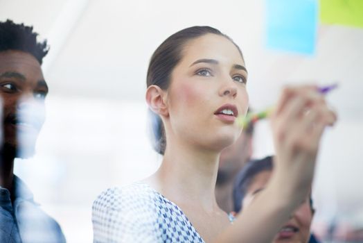 Cropped shot of an attractive young businesswoman making notes on a glass wall while her colleagues look on.