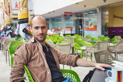 Adult man with casual clothing sitting at a table outside a coffee shop smiling and looking at the camera