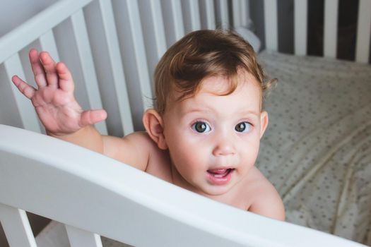 Cute baby with curly blonde hair standing in a cot, looking and waving at the camera