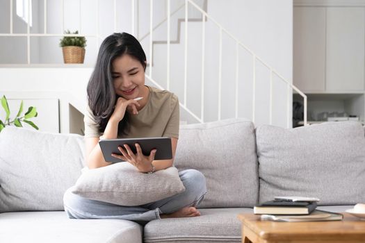 Attractive smiling young asian woman relaxing on a leather couch at home, working on laptop computer..