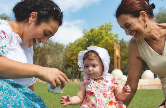 A happy family of 3 generations, baby, mother and grandmother sitting on the grass and playing outdoors in a public park