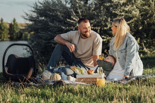 Happy Couple with Their Toddler Son Having a Picnic Outdoors by Lake, Young Family Weekend Concept