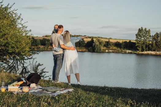 Happy Young Family, Mother and Father with Their Little Son Having Picnic Outdoors Near Lake at Sunset