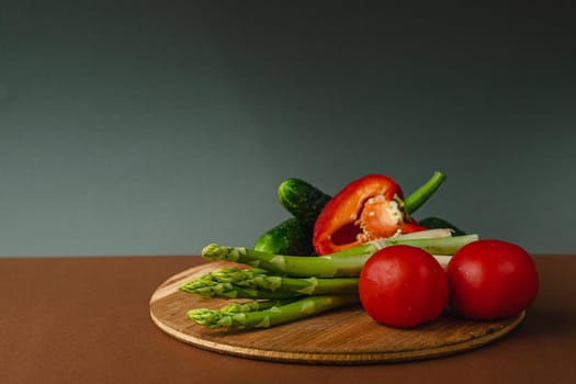 Vegetables lie on a wooden board: tomatoes, asparagus, cucumbers, red bell peppers. brown, dark gray background. place for text