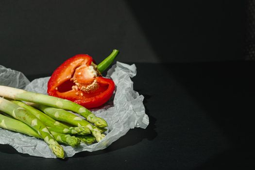 fresh green asparagus and half of red sweet, large pepper lie on white parchment on a dark gray background. proper nutrition. Lifestyle. place for text